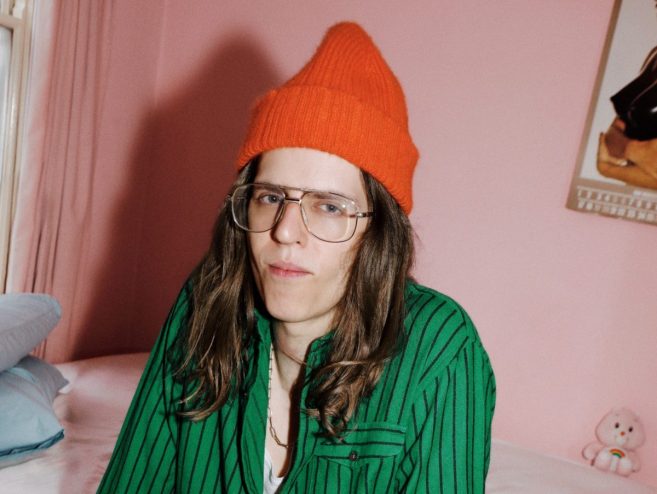 Bunny Kinney sits on a bed in a pink bedroom wearting a bright orange beanie and striped green shirt