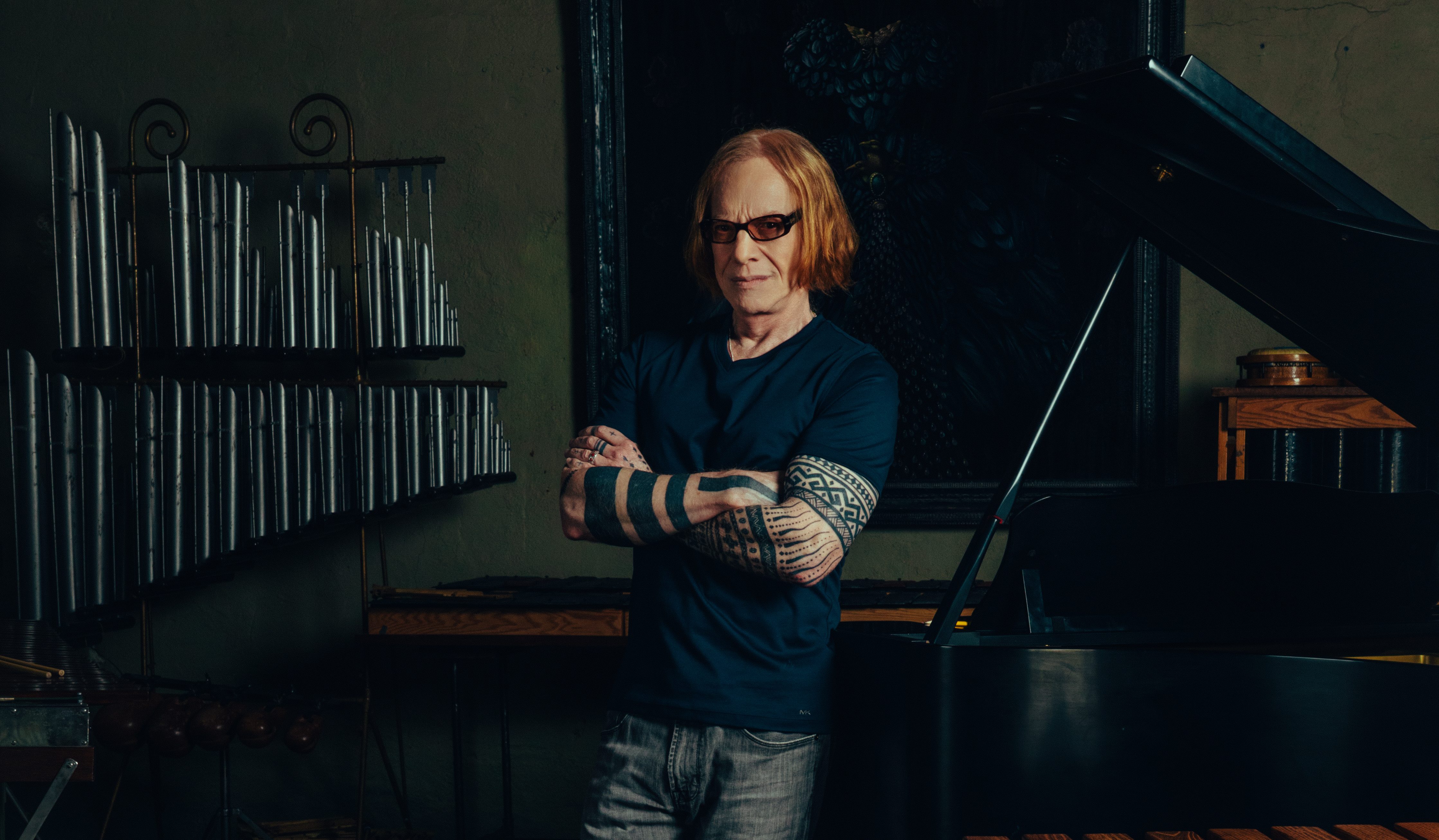 Danny Elfman stands against the piano in his studio wearing a blue shirt and sunglasses