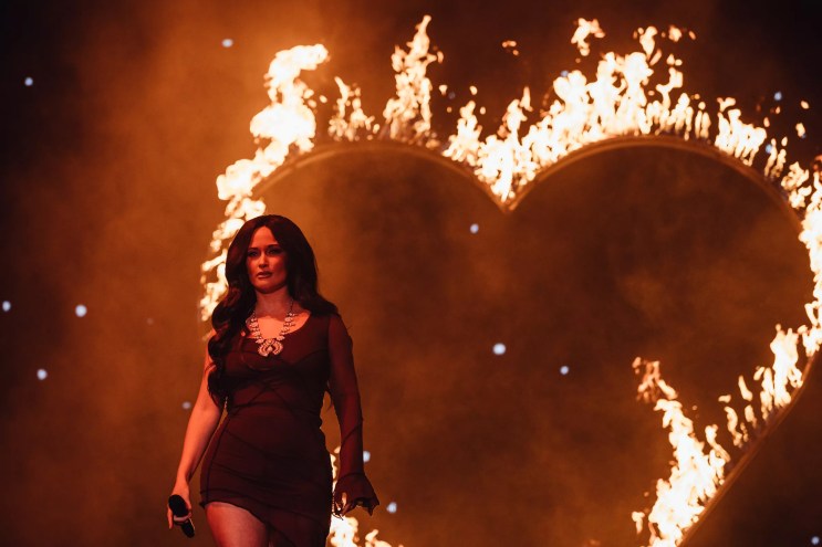 Kacey Musgraves stands on stage in a black dress in front of the shape of a heart on fire