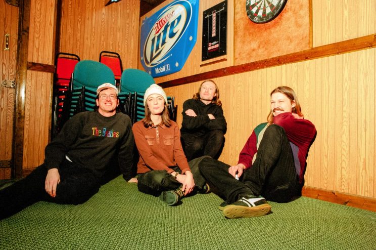indie rock band ratboys sits on green carpet surrounded by wood-paneled walls with a blue miller lite sign and dart board hanging above