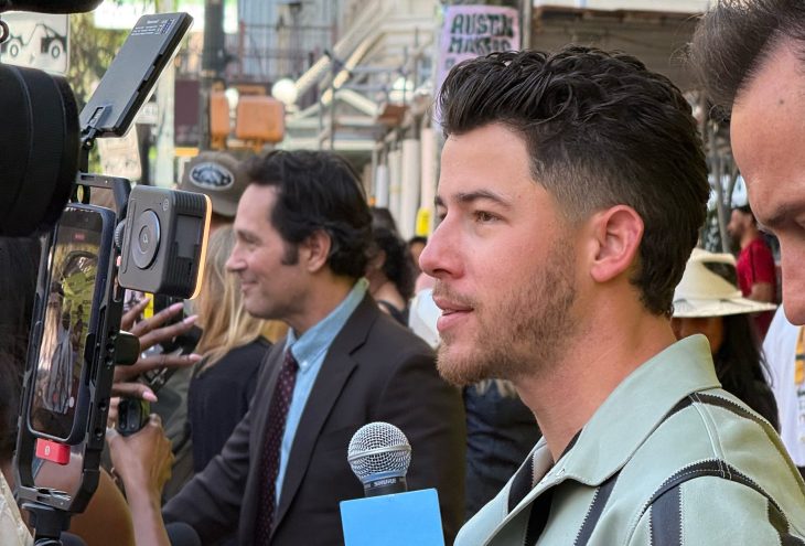 a side profile of nick Jonas wearing a mint green and black striped jacket as Paul Rudd speaks to reporters in the background