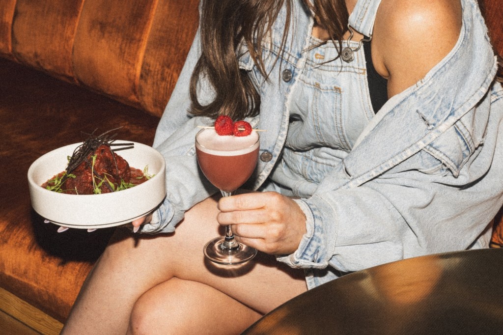 Woman at Apothecary holds a plate of meatballs and a French kiss cocktail with raspberry vanilla vodka, pineapple oleo, egg white, lemon and raspberries.