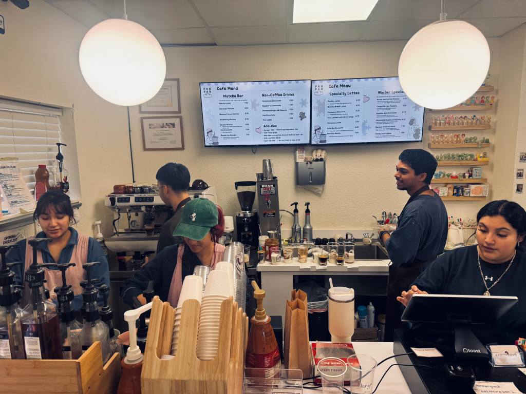 a busy cafe counter with five baristas working the cash register and making drinks.