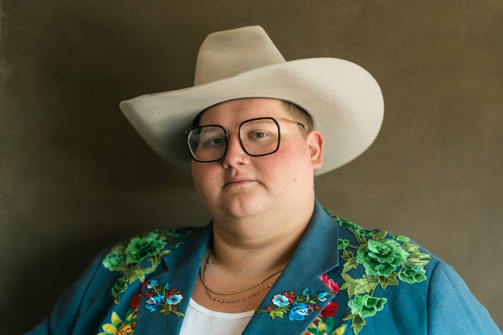 joshua ray walker looks into the camera wearing a white cowboy hat, glassses and an embroidered turquoise blazer