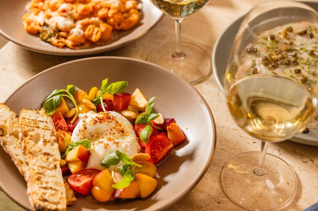 Burrata served with colorful tomatoes, sliced stone fruit, basil, and toasted bread, accompanied by glasses of white wine.