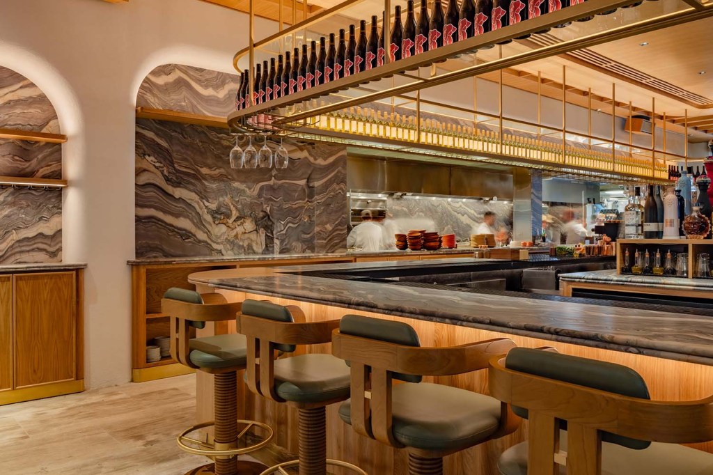 Elegant restaurant bar with marble counters, wood cabinetry, brass shelving, and rows of wine bottles displayed overhead.