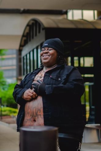 Rapper BigXthaPlug smiling in front of building with shirt unbuttoned wearing a beanie